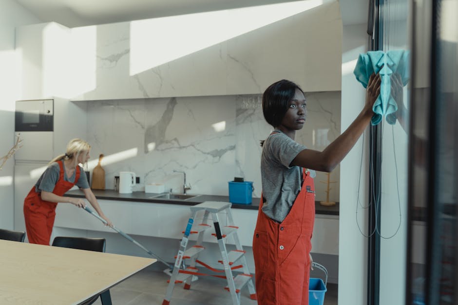 Two professional cleaners in red uniforms are engaged in surface cleaning within a modern kitchen. The cleaner in the foreground is wiping a glass surface or window with a blue cloth, focusing on removing dust or smudges, while the other is using a mop to clean the tiled or wooden floor. The kitchen features a sleek design with a marble backsplash, stainless steel appliances, and a black countertop, all appearing spotless and well-maintained. Natural light streams into the room, highlighting the polished surfaces and clean environment, emphasizing thorough domestic cleaning by Ilford Cleaners as part of their high road house cleaning services.