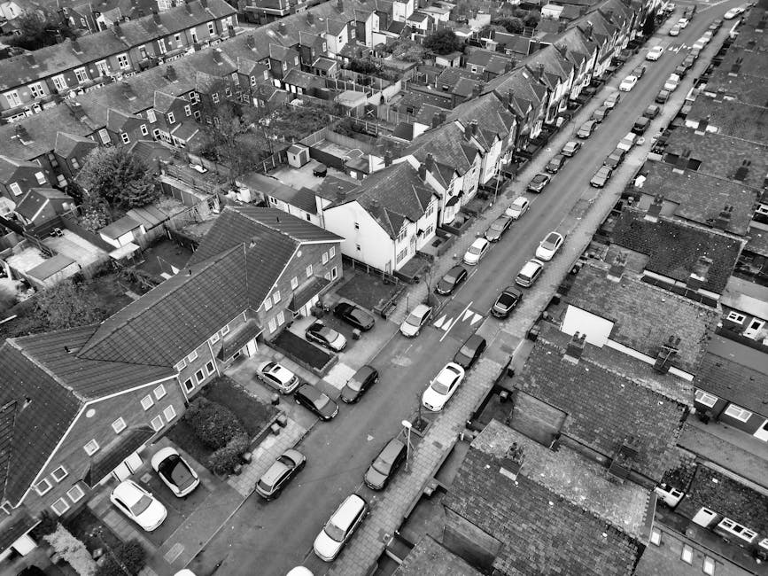 An aerial black-and-white image of a residential street showing rows of houses with pitched roofs, some with dormer windows, and small front gardens. The street is lined with parked cars on both sides, including sedans and hatchbacks, with a few visible road markings and a pedestrian crossing. The houses are primarily constructed of brick and wood, with tiled roofs and small windows. The overall scene appears clean and orderly, with no visible debris or clutter. Street lamps are present along the sidewalk, and the lighting suggests daytime with even illumination across the scene. The image conveys a quiet neighborhood atmosphere, captured by Ilford Cleaners as part of their residential cleaning and maintenance content, emphasizing the importance of well-maintained outdoor environments.