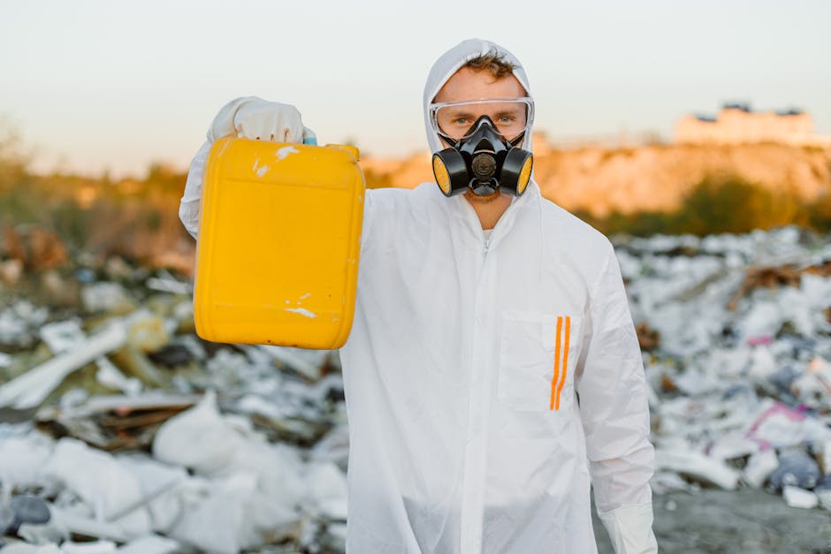 A person dressed in white protective coveralls, wearing a respirator mask and goggles, stands outdoors at a waste disposal site with scattered debris and plastic waste on the ground. The individual is