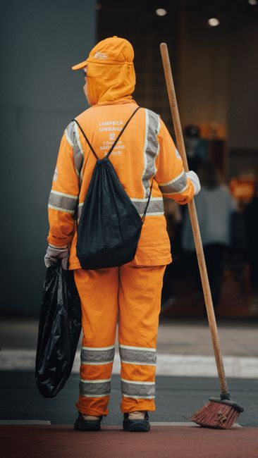A sanitation worker dressed in a bright green uniform and matching cap is leaning over a large green outdoor waste bin, which contains various cleaning tools such as a broom and a mop. The worker appe