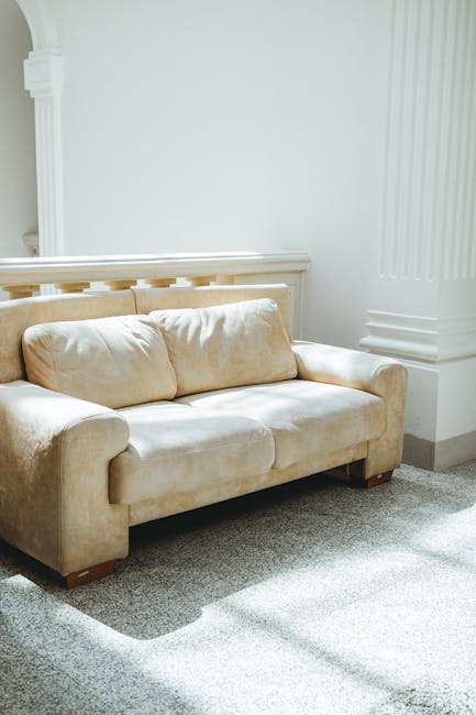 A beige fabric upholstered sofa positioned against a white wall with decorative molding in a well-lit living room. The sofa appears clean and plush, with two large seat cushions and matching back cush
